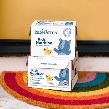 Two cases of Kate Farms Kid's Nutrition Shake, Vanilla, from Kate Farms sit on a colorful rainbow doormat by a yellow wall. The boxes display a bear illustration along with product details.
