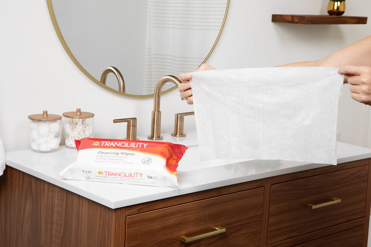 A person holds a large sensitive skin wipe above a bathroom sink, with a pack of Tranquility Cleansing Wipes with Aloe by Tranquility on the counter next to jars of cotton balls and swabs.