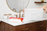 A person holds a large sensitive skin wipe above a bathroom sink, with a pack of Tranquility Cleansing Wipes with Aloe by Tranquility on the counter next to jars of cotton balls and swabs.