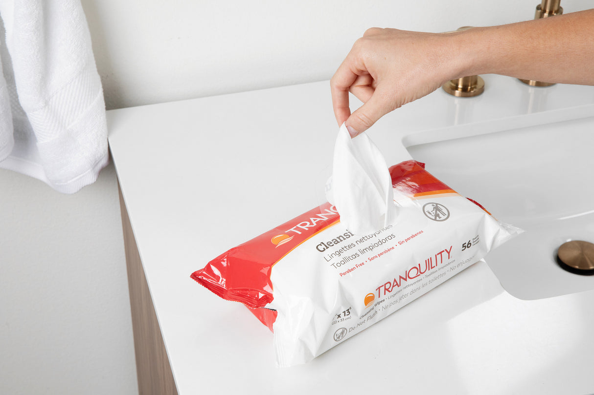 A hand pulls a Tranquility Cleansing Wipe with Aloe from its pack, placed on a white bathroom sink beside a towel and faucet.