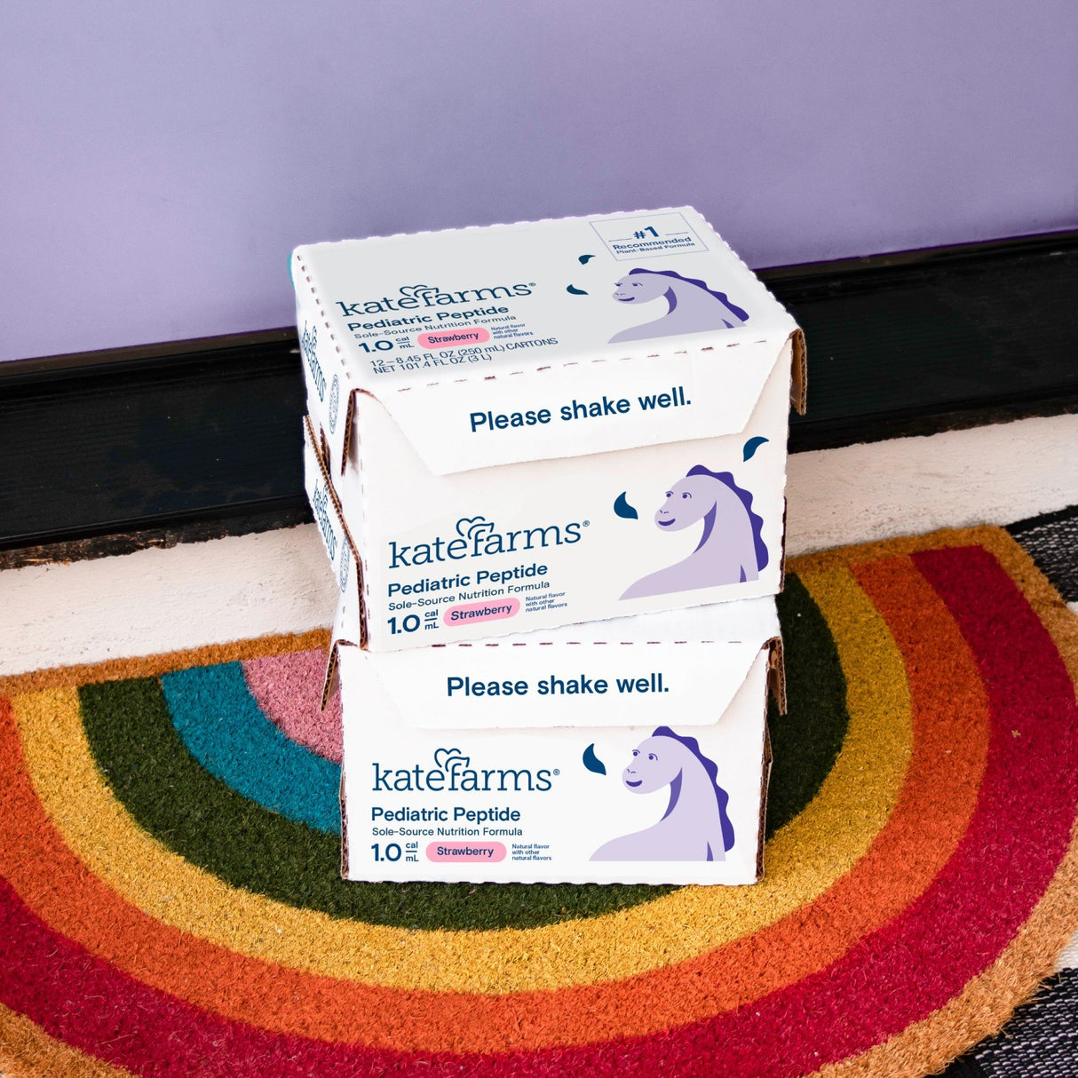 Two boxes of Kate Farms Pediatric Peptide Formula 1.0, Strawberry, sit on a rainbow doormat by a purple wall. The plant-based formula boxes feature a unicorn and the text "Please shake well.