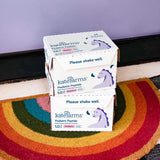 Two boxes of Kate Farms Pediatric Peptide Formula 1.0, Strawberry, sit on a rainbow doormat by a purple wall. The plant-based formula boxes feature a unicorn and the text "Please shake well.