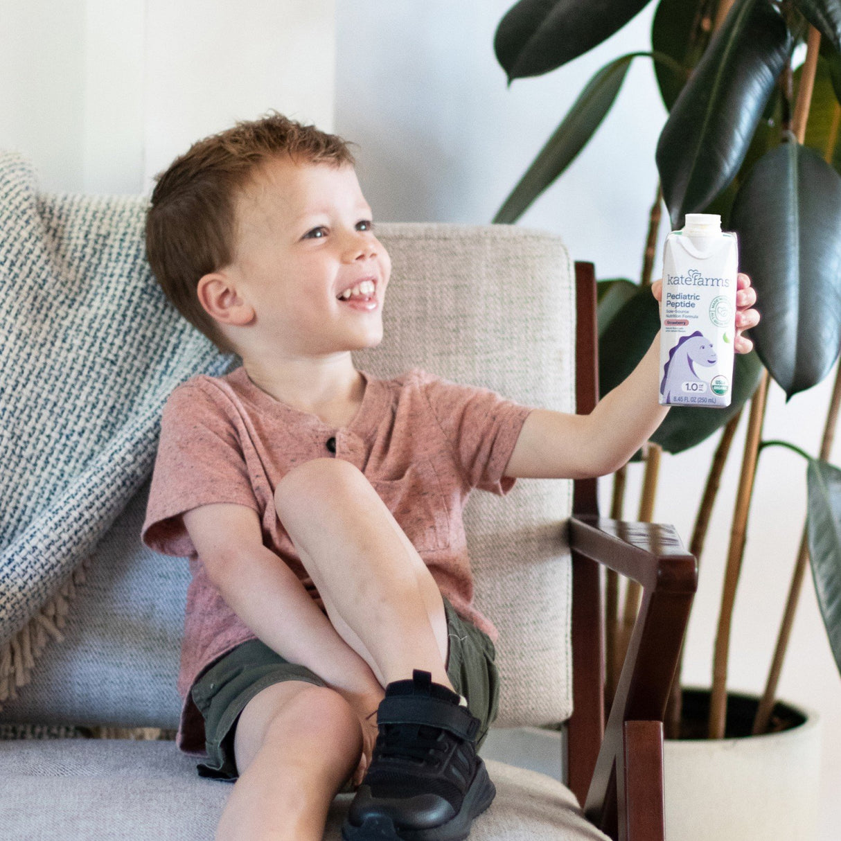 A young boy smiles while sitting on a light chair, holding up a carton of Kate Farms Pediatric Peptide Formula 1.0, Strawberry. The plant-based nutrition drink from Kate Farms features a unicorn design, with a leafy plant and blanket in the background.