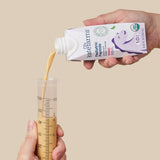 A hand pours Kate Farms Pediatric Peptide Formula 1.0, Strawberry from a Kate Farms carton into a large medical syringe held by another hand, illustrating tube feeding against a plain beige background.