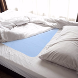 A bed with white sheets and a pillow features a NorthShore Champion XD Washable Underpad placed below the fitted sheet, covering the middle section. Sunlight streams through a window in the background.