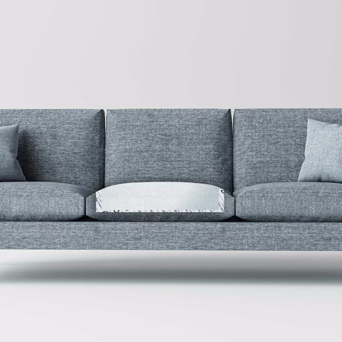 A modern gray sofa with three cushions has two gray throw pillows on each end, and the middle cushion is flipped to reveal a NorthShore MagicSorb Super-Absorbent Disposable Underpad, showing its lighter, odor-absorbing, waterproof underside. Plain white background.
