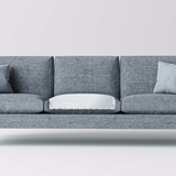 A modern gray sofa with three cushions has two gray throw pillows on each end, and the middle cushion is flipped to reveal a NorthShore MagicSorb Super-Absorbent Disposable Underpad, showing its lighter, odor-absorbing, waterproof underside. Plain white background.